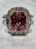 A cushion-cut pink tourmaline ring with a halo of small diamonds and baguette cut side stones, set in a silver-toned metal on a light marble surface.
