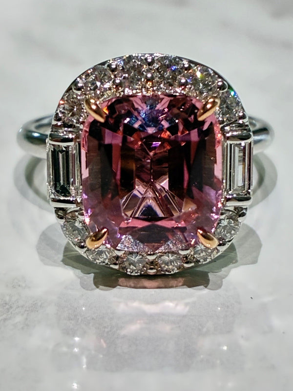 A cushion-cut pink tourmaline ring with a halo of small diamonds and baguette cut side stones, set in a silver-toned metal on a light marble surface.