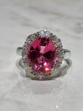 A white gold ring featuring a large oval pink Tourmaline surrounded by a halo of small marquise shaped diamonds, displayed on a light marble surface.