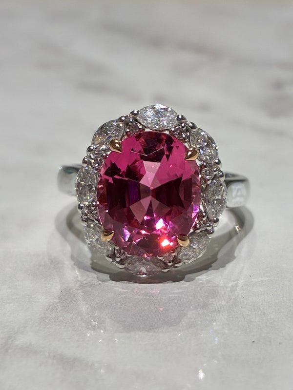 A white gold ring featuring a large oval pink Tourmaline surrounded by a halo of small marquise shaped diamonds, displayed on a light marble surface.