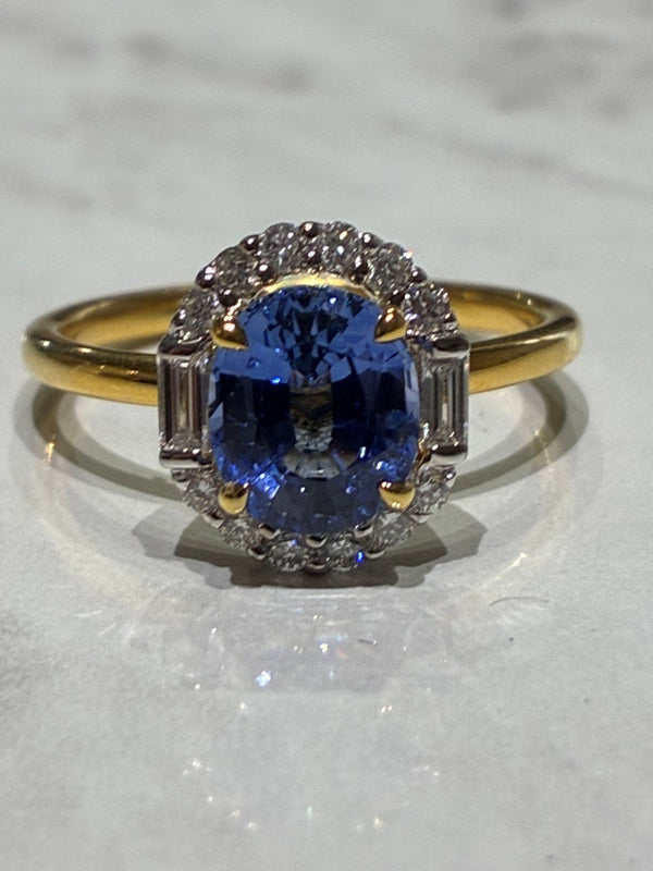 A yellow gold ring with a central oval-cut sapphire surrounded by small diamonds and baguette side stones, displayed on a light marble surface.