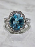 A white gold halo ring featuring a large oval-cut aquamarine, accented with small pavé-set stones along the halo and split shank, displayed on a light marble surface.