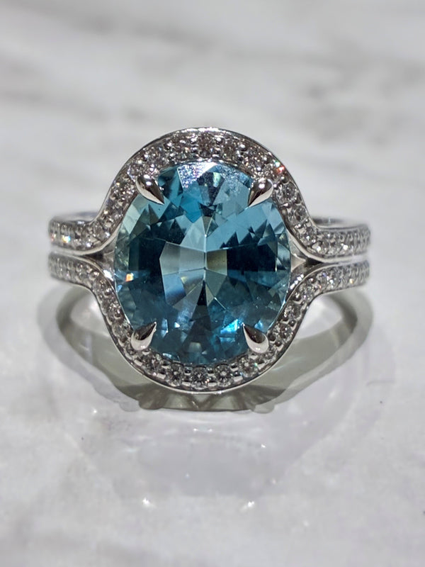 A white gold halo ring featuring a large oval-cut aquamarine, accented with small pavé-set stones along the halo and split shank, displayed on a light marble surface.