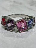 A white gold ring featuring five colorful spinel in a row: red, lavender, pink, blue, and purple, displayed on a light marble surface.