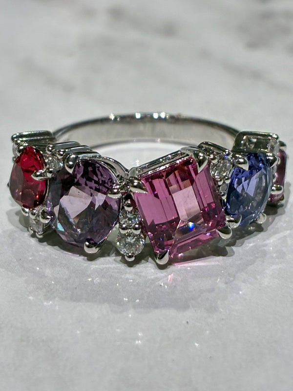 A white gold ring featuring five colorful spinel in a row: red, lavender, pink, blue, and purple, displayed on a light marble surface.