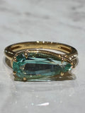A gold ring featuring a pear shape light green emerald, displayed on a white marble surface.