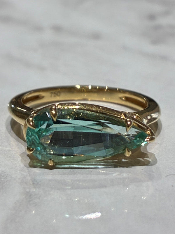 A gold ring featuring a pear shape light green emerald, displayed on a white marble surface.