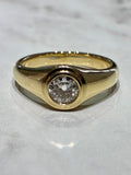 A yellow gold signet-style ring with a single old cut round diamond, resting on a light gray marble surface.
