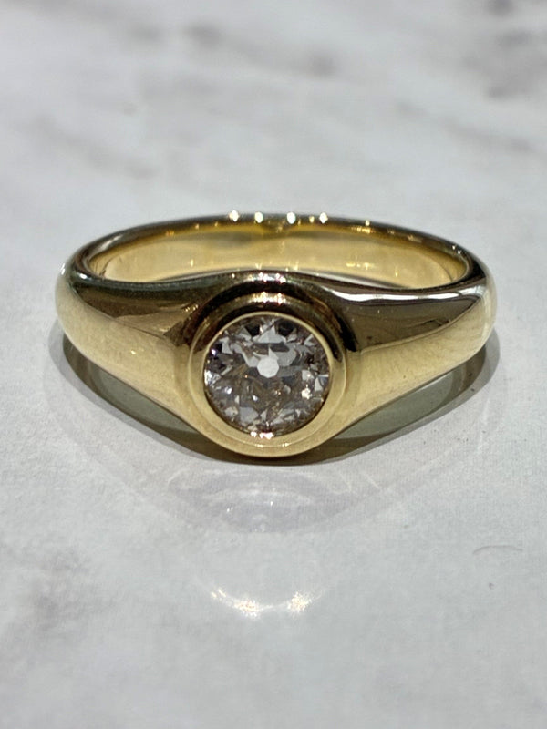 A yellow gold signet-style ring with a single old cut round diamond, resting on a light gray marble surface.